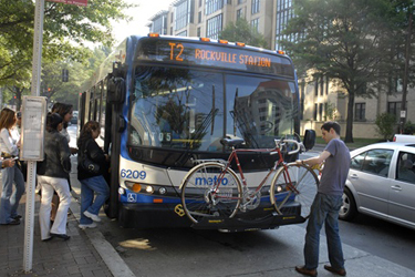 A DC Metro bus taking on passengers while a cyclist puts his bicycle on the front of the bus.